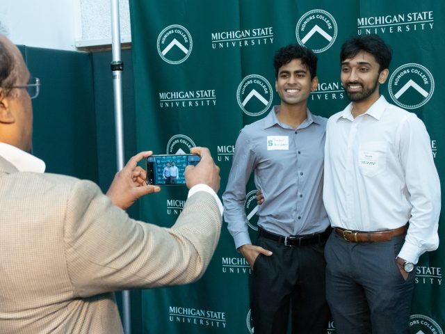 Decorative image of two graduates at an HC Graduation Reception smiling while having their photo taken by a person in a brown jacket in front of a green Honors College backdrop