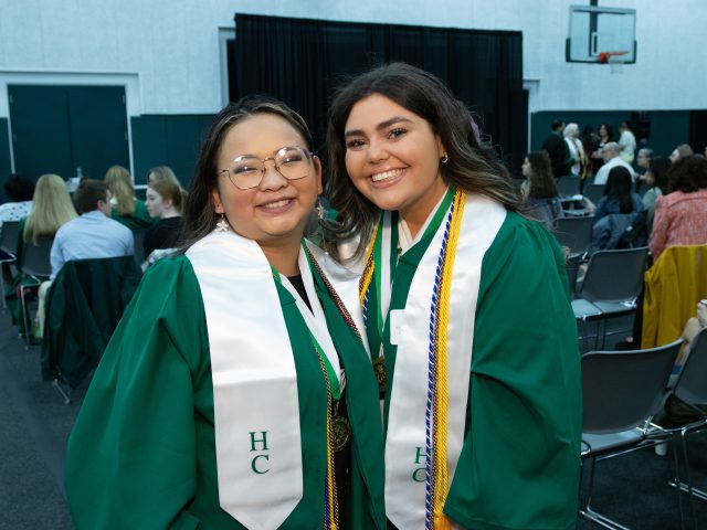 Decorative image of two HC graduates smiling at the camera during an HC Graduation Reception wearing their green graduation gowns and the white HC stole