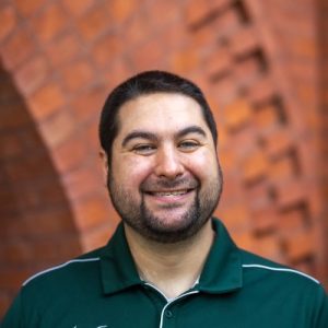 Headshot of Andrew Abad; person smiling at camera in front of brick building wearing green polo