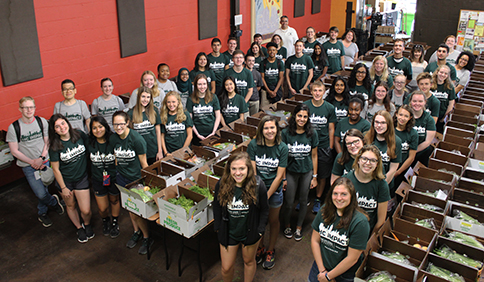 2018 IMPACT participants and staff smiling at the camera wearing program T-shirts in front of boxes of produce at the Allen Neighborhood Center