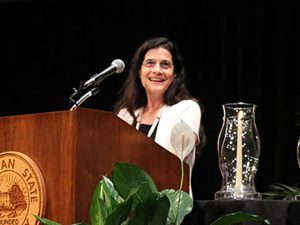 Photo of Jeanne Wald standing behind a podium bearing the MSU seal wearing a white shirt with a lily plant and glass vase with candle next to the podium