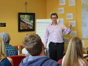 Decorative image of John Waller (person standing in plaid white shirt) teaching a class of students who are seated and facing the front of the classroom. Classroom pictured has yellow walls and various items hung on the wall and a white easel behind John Waller. 