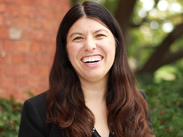 Headshot of Melanie Brender; person smiling at camera in front of brick building wearing a polka dot shirt and black blazer with shoulder-length brown hair