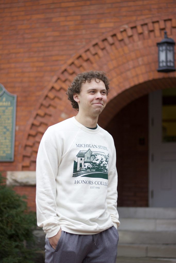 A man in an off-white MSU Honors College sweater smiles in front of a red brick building