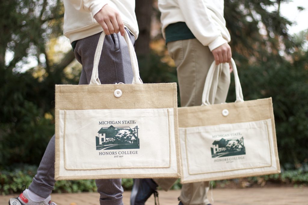 Two people are walking by, and the image is a close-up of the matching jute and canvas tote bag each person is carrying