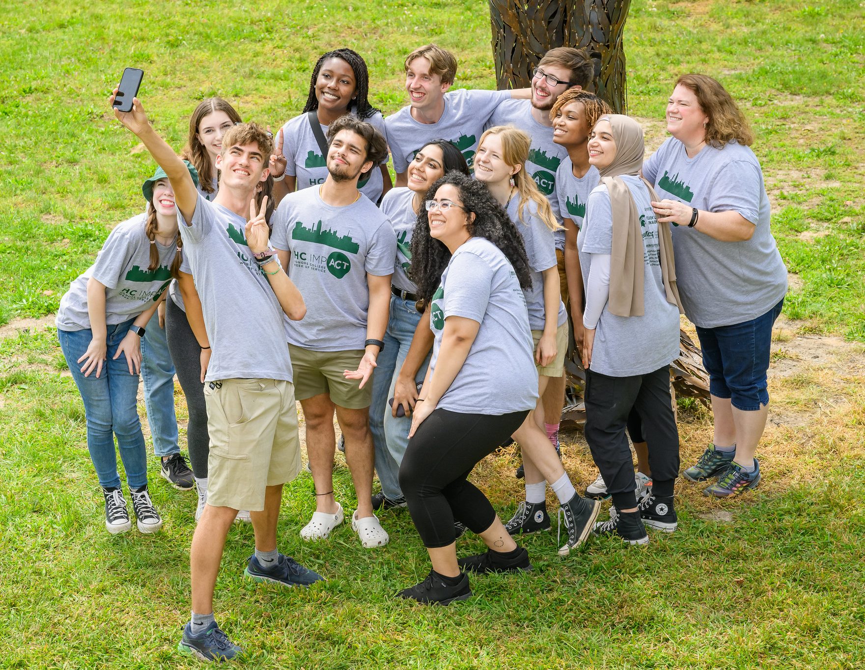 A group of smiling students in grey and forest green HC IMPACT shirts stand on the lawn to take a group selfie