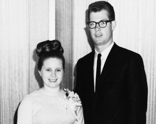 A black-and-white photo of a man and a woman in formal attire. The man as a skinny black tie, glasses, and suit. The woman is in a lighter sleeveless dress.