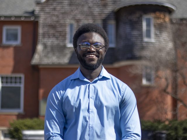 A smiling student in wire-rimmed classes and a blue collared shirt stands in front of a red brick building with wooden shingles.