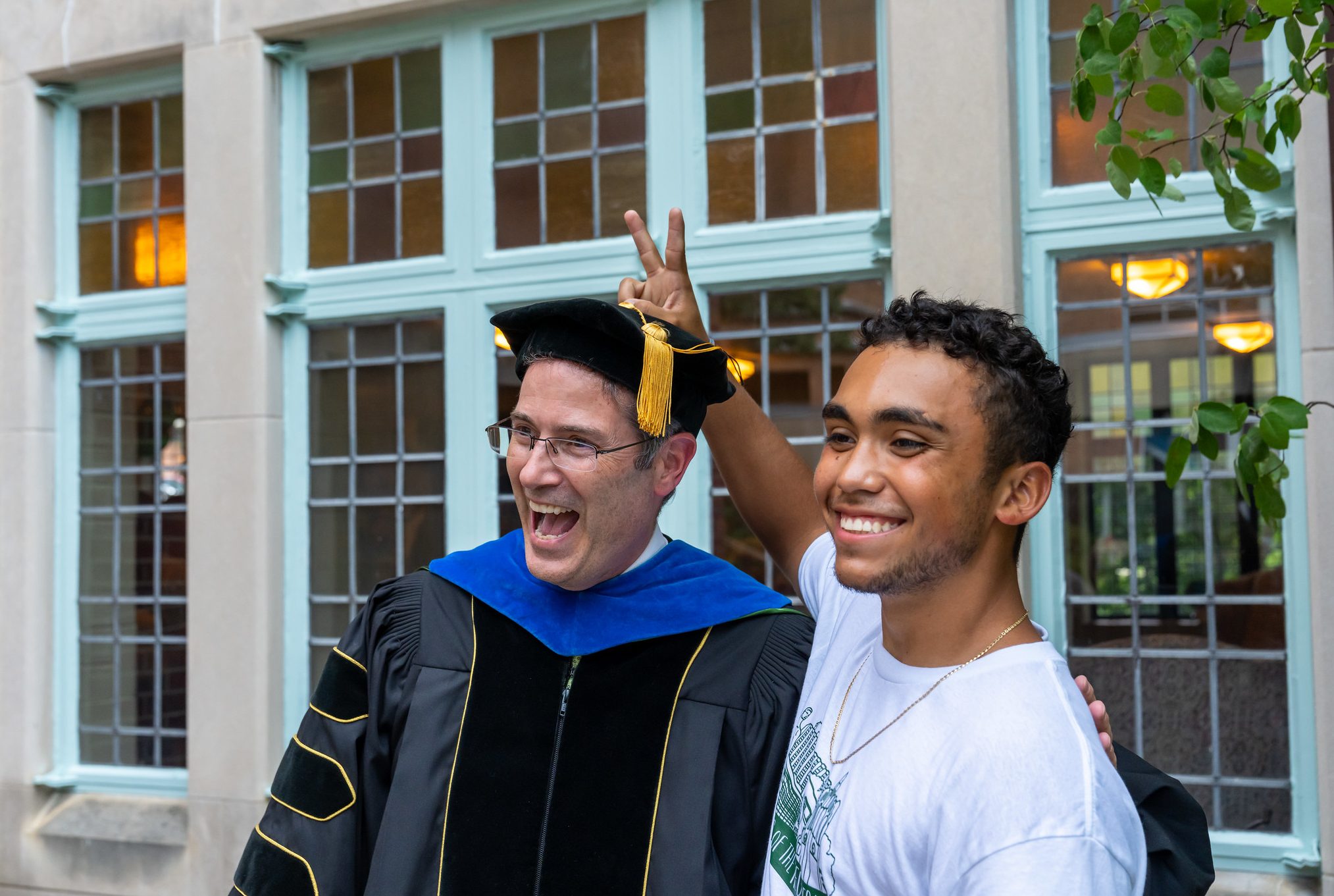 A student in a white t-shirt holds two fingers behind the head of a man in black PhD graduation robes.