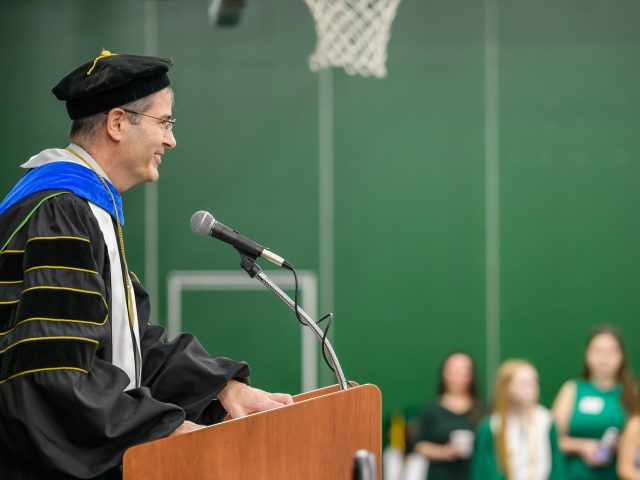 A man in black PhD graduation robes standing at a podium with a microphone