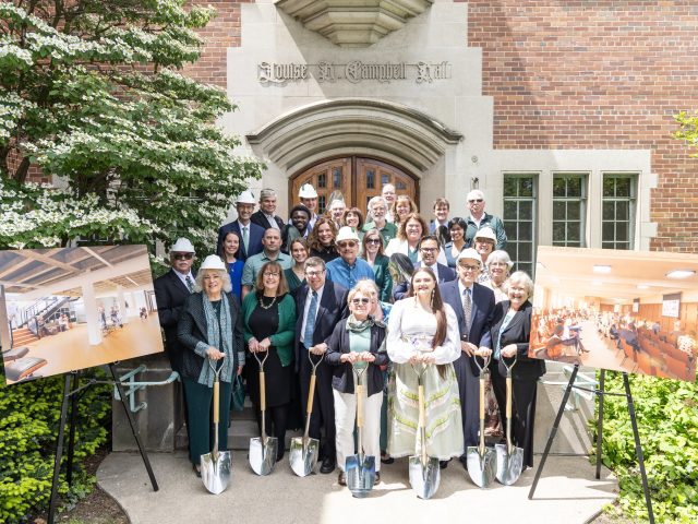 A group of people stands on the steps of Michigan State University's Campbell Hall. Some of them are in white hard hats, while others hold gleaming silver shovels. On each side of the stairs is foam core poster on a easel with renovation renderings.