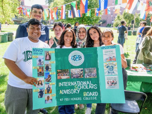 A group of seven college students hold up a green poster board that reads 
