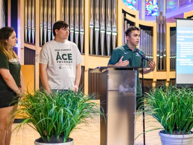 Three students stand up at a podium, speaking to an unseen audience. One student's shirt reads 