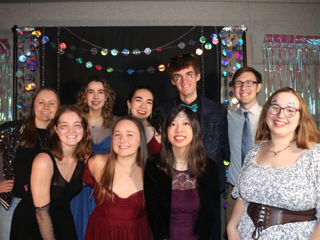 A group of students in formal-wear smiling at the camera, with a glittery, festive backdrop in the background.