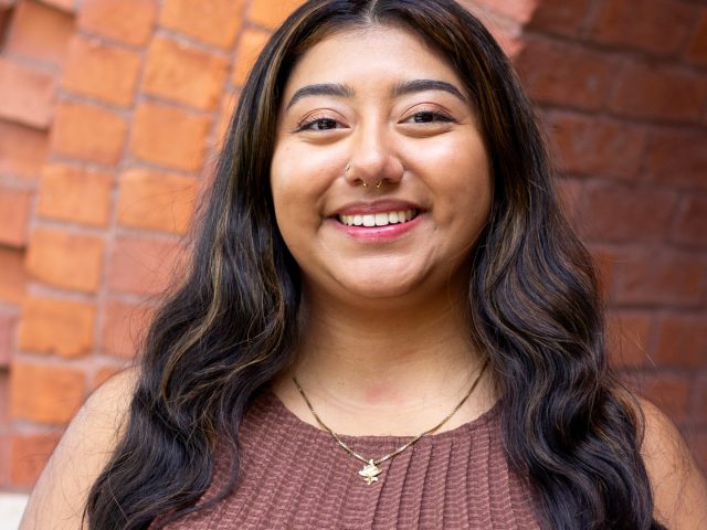 Honors College Executive Assistant Alyssa Briones smiling in front of the red brick entrance to Eustace-Cole Hall.