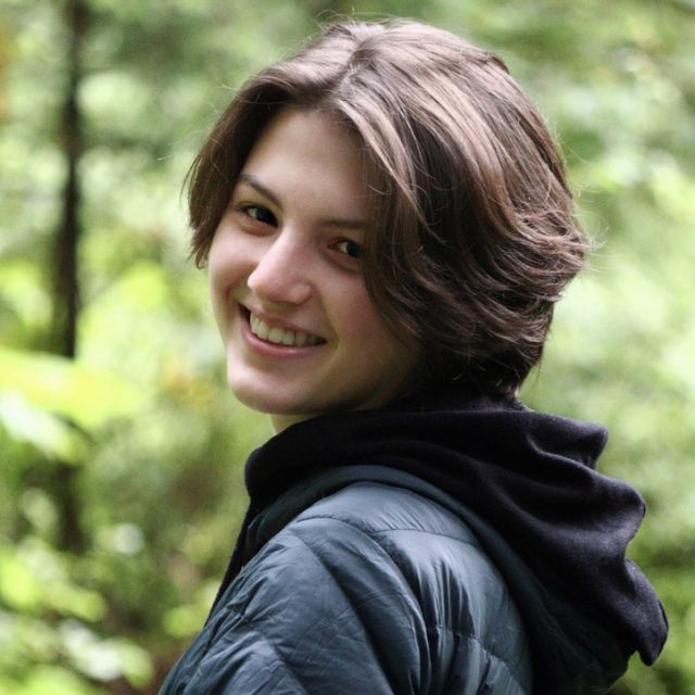 Photo of Annie Walton in a black jacket looking over the shoulder at the camera, smiling. Lush forest is in the background.