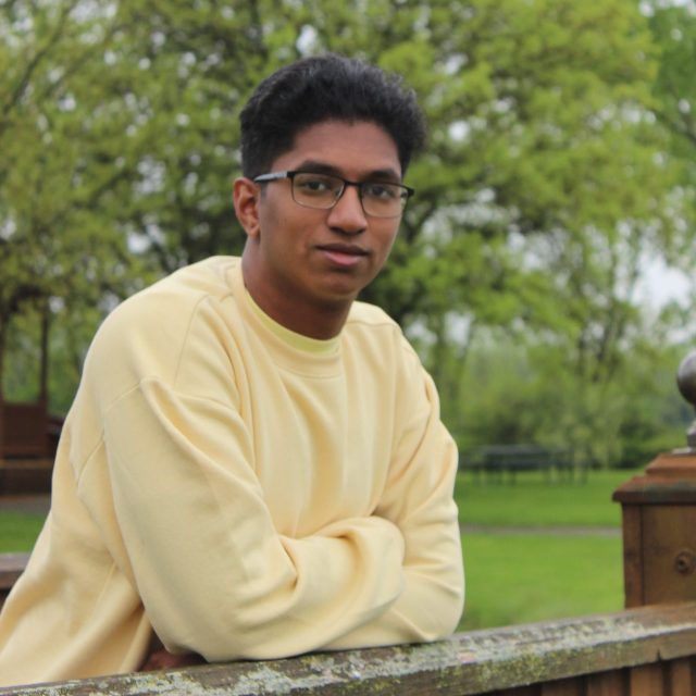 Photo of Guatam Kiran in a yellow sweater, leaning on a wooden ledge. Grass and trees are in the background.