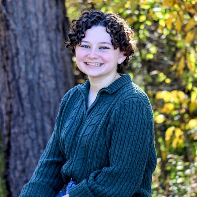 Photo of Julia Bolash in a green sweater and jeans, sitting on a log in the woods and smiling.