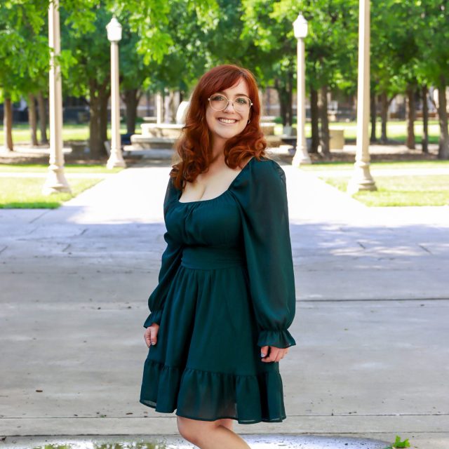 Photo of Katherine Dyal in a long-sleeve dress, smiling. Pavement, lamp posts, and trees are in the background.