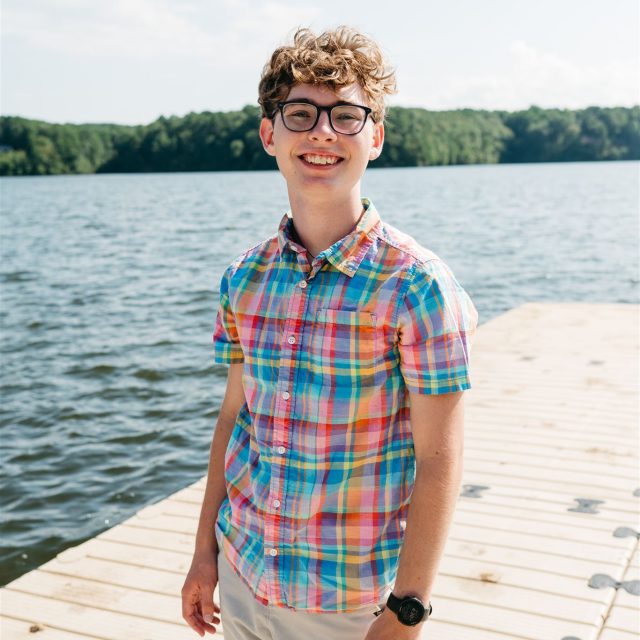Photo of Zachary Bigelow standing on a dock in a colorful plaid shirt, smiling. There is a body of water in the background.
