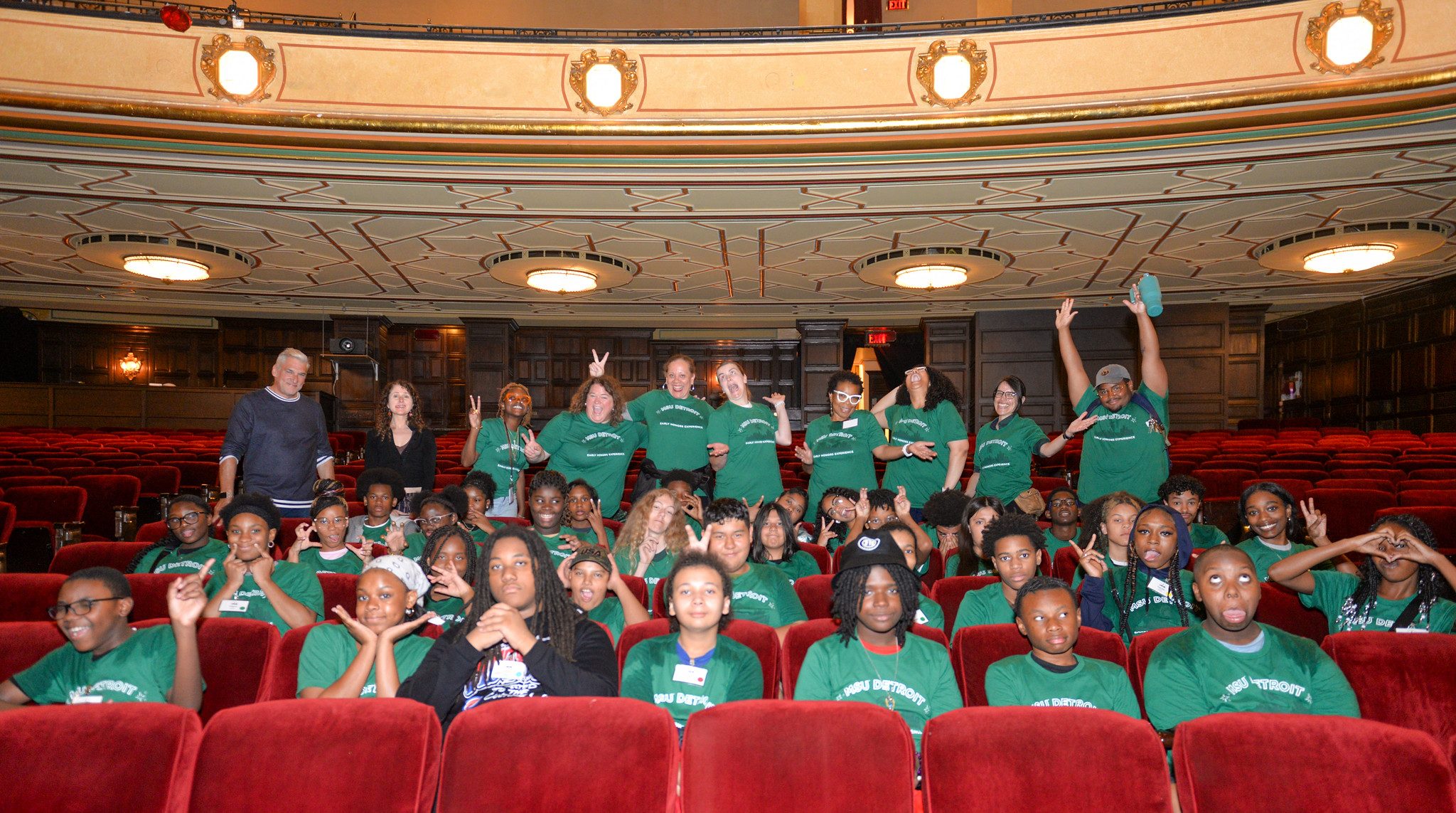 A group of students and staff members in multiple rows of red Detroit Opera House seats. Some are standing, some are sitting, and many are posing with fun expressions, peace sign gestures, and smiles.