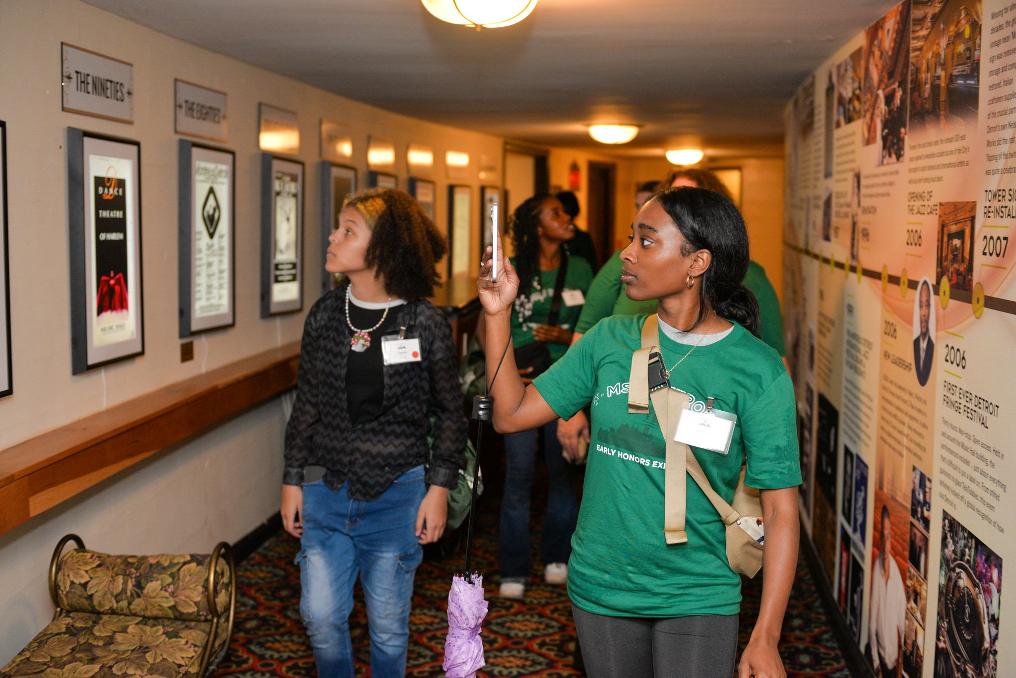 Students walk down a hallway where the walls are covered with music history information. One student holds out her phone to take a photo.