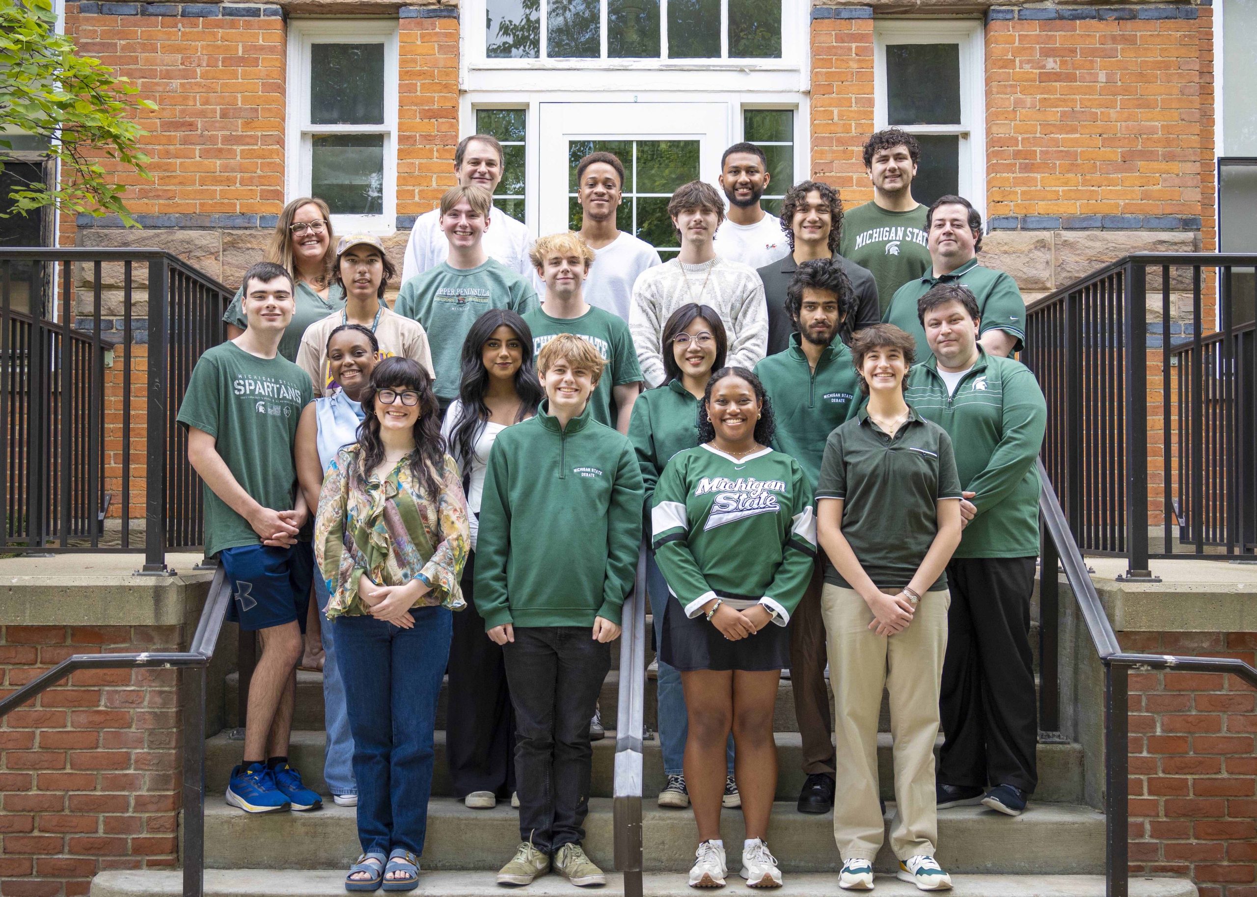 The MSU Debate Team stands in a group on the steps of Linton Hall. Most of them are wearing green or white shirts.