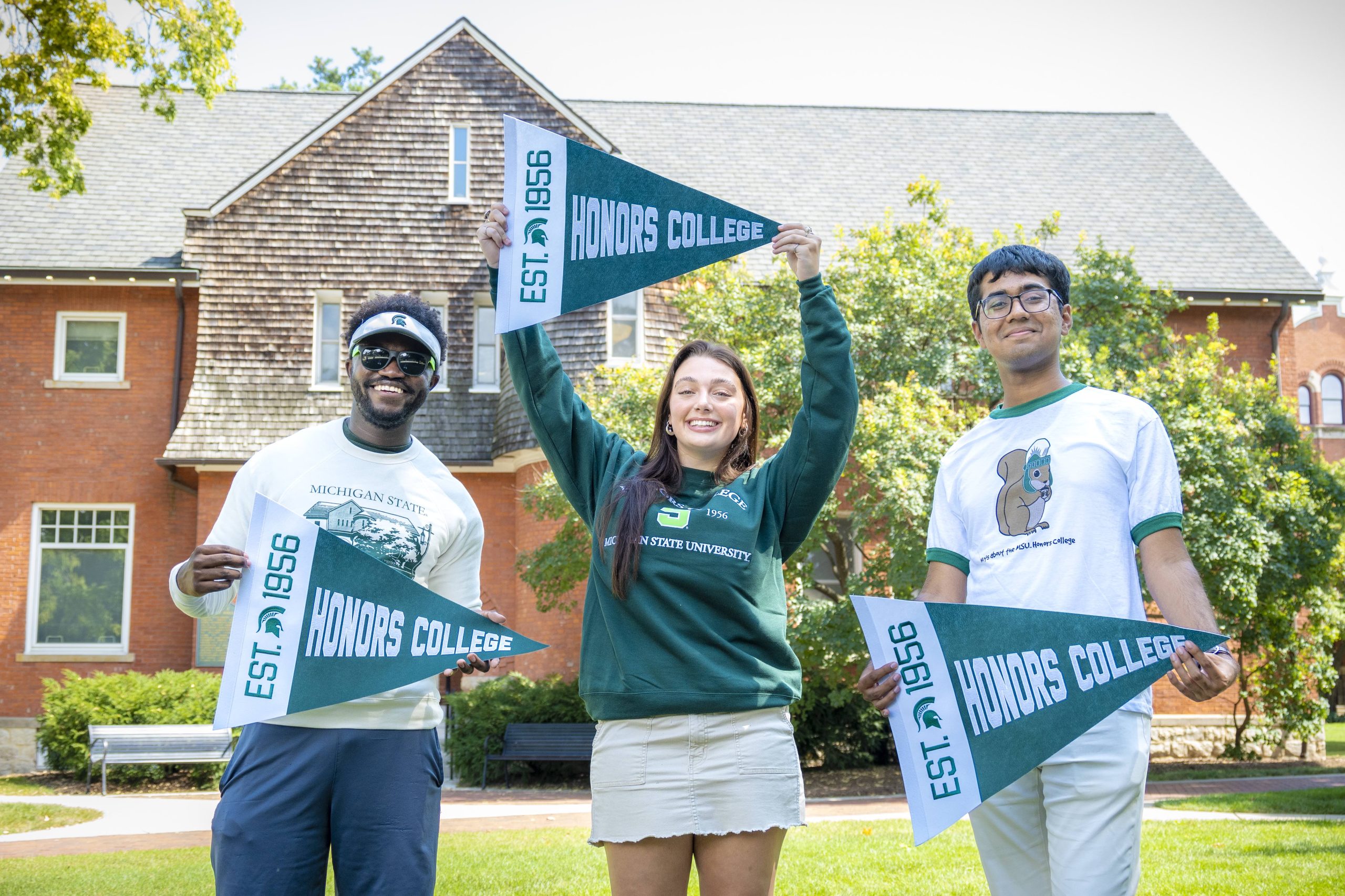 Three students standing outdoors in front of a Eustace-Cole Hall, smiling and holding green and white MSU Honors College pennants with 'EST. 1956' printed on them. The students are wearing MSU Honors College apparel.