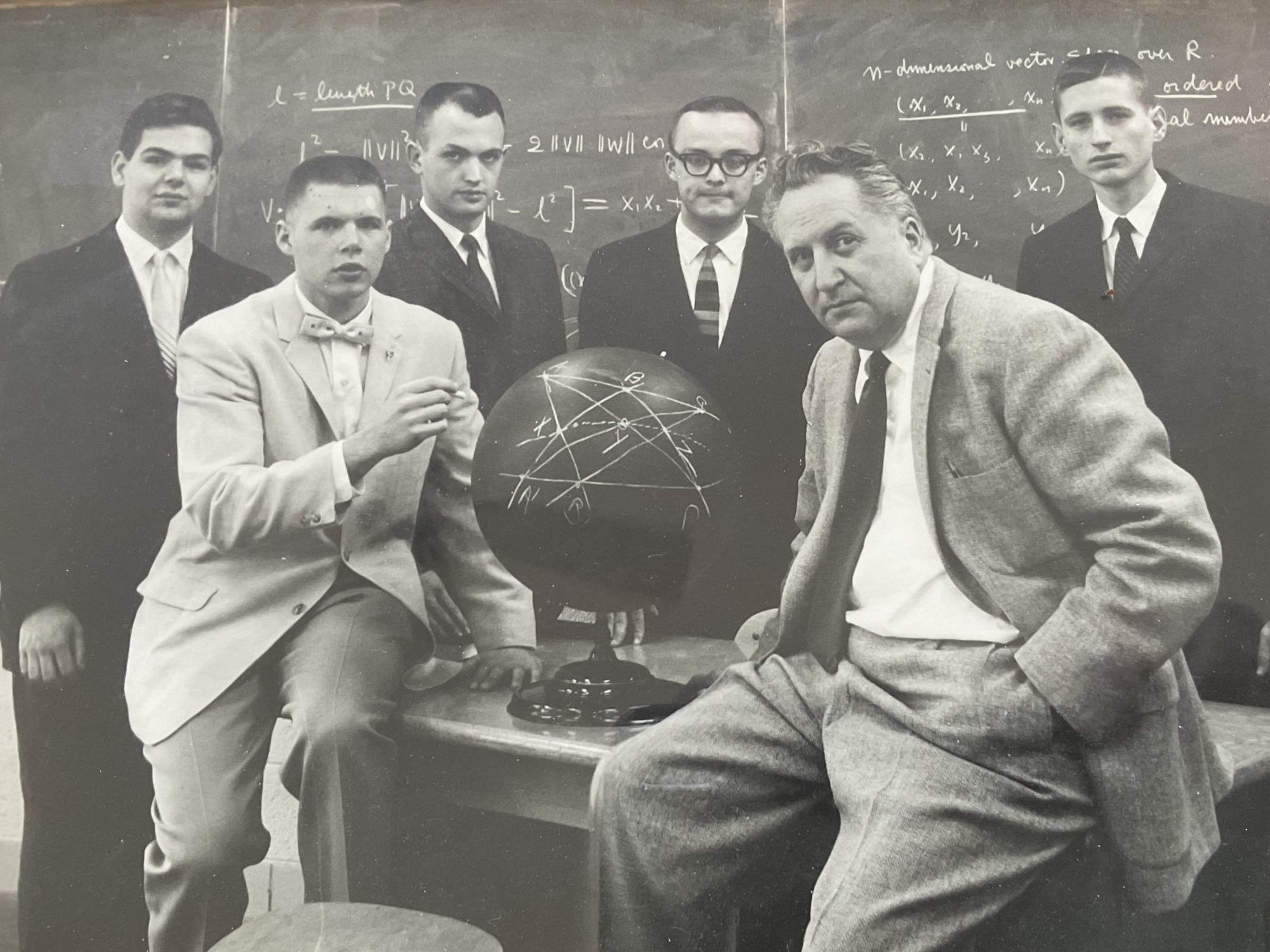 A black-and-white photo of six men in formal attire, standing around a globe with mathematical equations drawn on it. Ted Petrie, wearing a light-colored suit and a bowtie, is seated on the table beside the globe, pointing at it. In the background is a chalkboard filled with mathematical formulas.