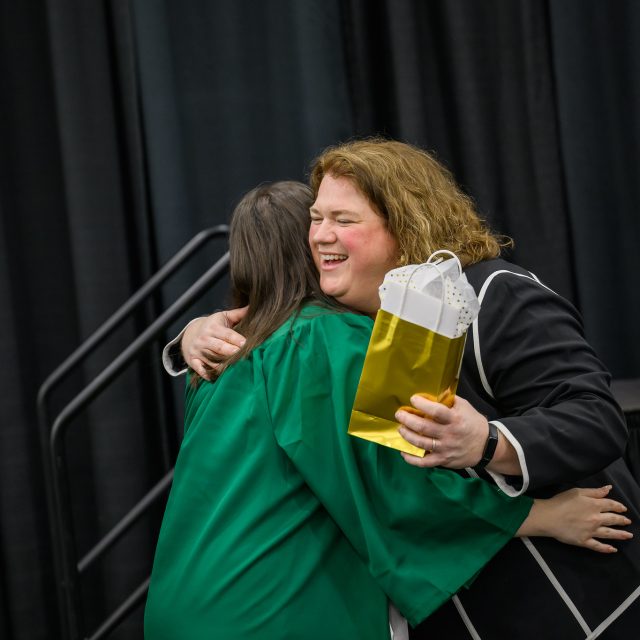 Bess German, wearing a black jacket with white trim, joyfully hugs a student in a green graduation robe. In Bess