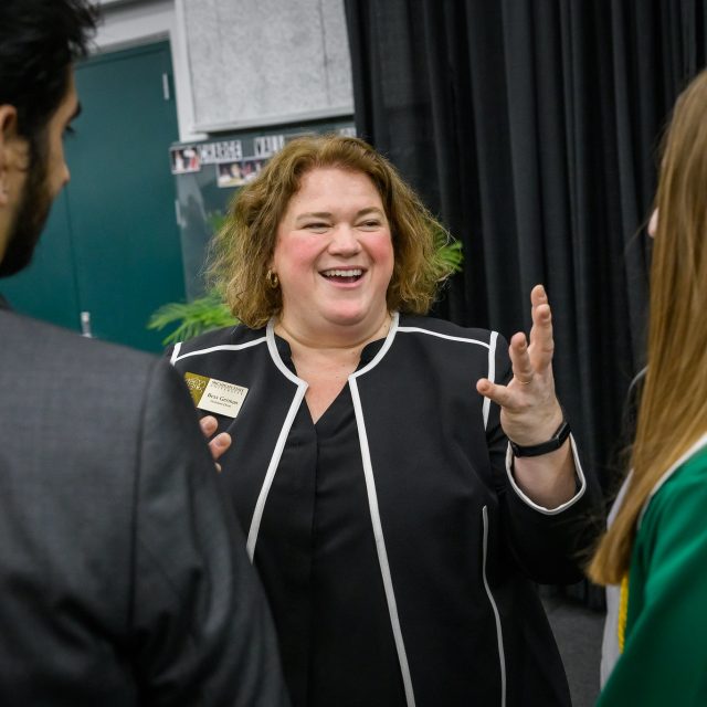 Bess German, wearing a black jacket with white trim, joyfully speaks with two students whose backs are to the camera. One student is in a green graduation cap and gown.