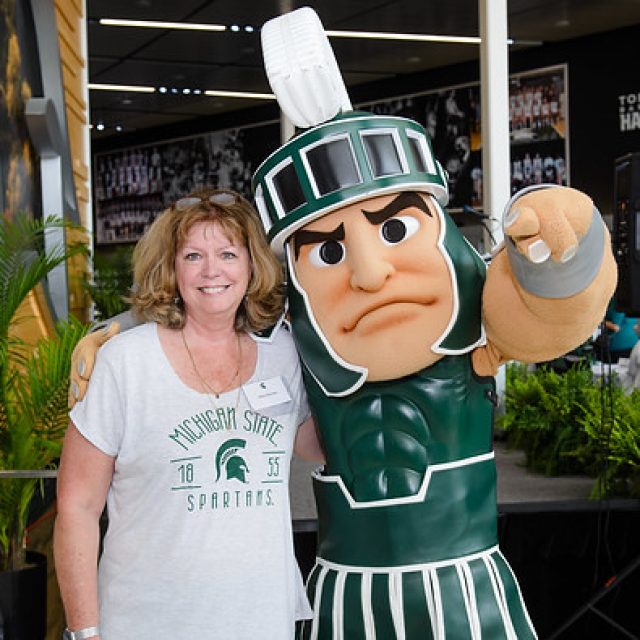 Molly Brennan, in a white shirt with a forest green Spartan helmet and text, poses with Sparty, who is pointing at the camera.