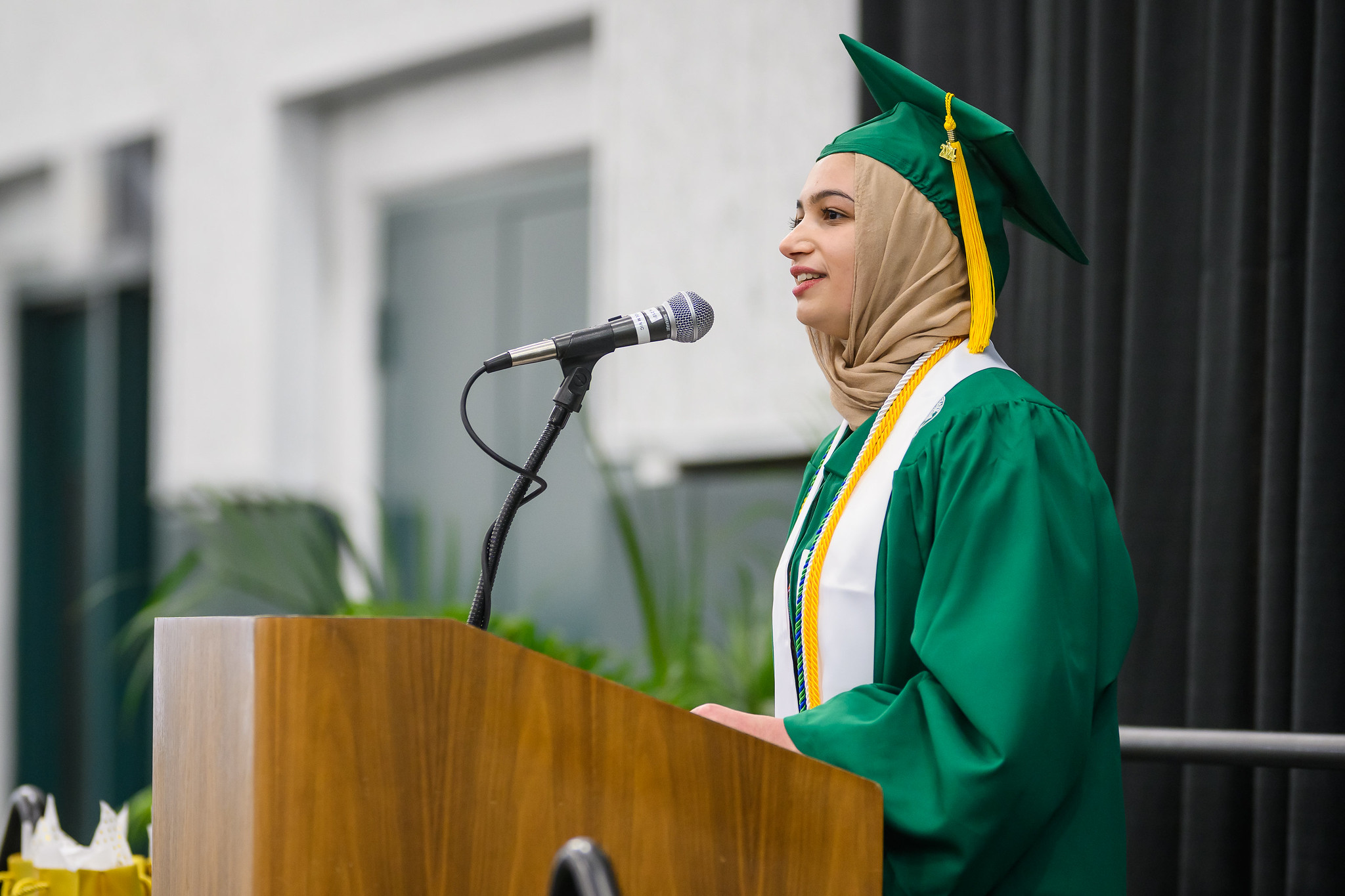 Honors College student Aya Abu-Zama speaks in a microphone at a wooden podium, wearing a green graduation cap and gown. Around Aya's neck is a white stole and three specially awarded graduation cords.