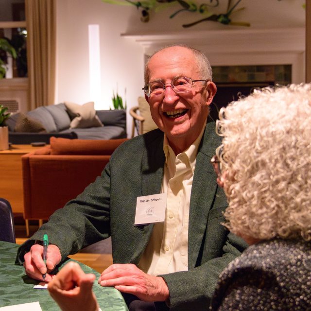 Bill Schoenl wears a white rectangular name tag, a forest green sports jacket, and a white collared shirt while sitting at a table and joyfully smiling at Cowles House. In the background are a fireplace, couches, and plants.