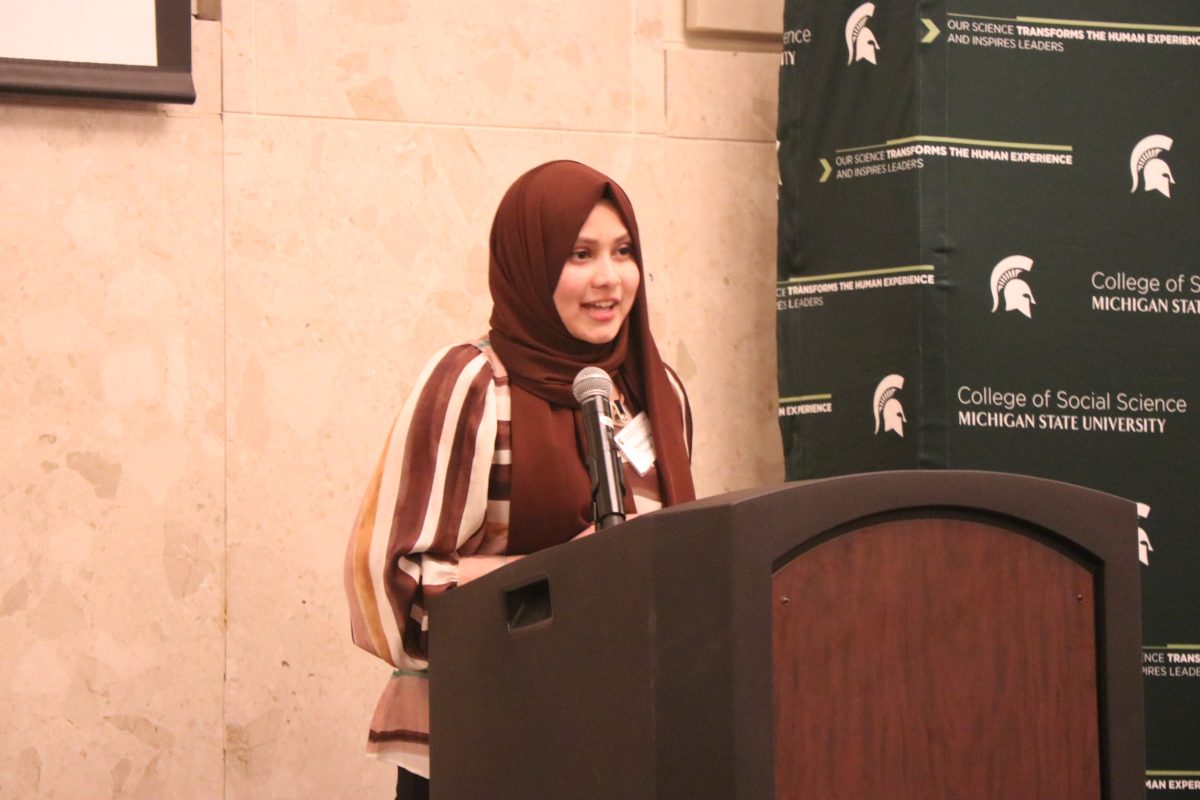 Sumaiya Imad speaking at the Women's Leadership Institute Recognition Ceremony. Imad is standing at a wooden podium with a microphone, wearing a brownish red hijab and striped blouse.