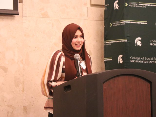 Sumaiya Imad speaking at the Women's Leadership Institute Recognition Ceremony. Imad is standing at a wooden podium with a microphone, wearing a brownish red hijab and striped blouse.