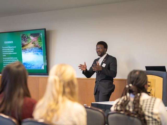 An Honors student presents research poster board to a group of people in the lounge of MSU Union, second floor.