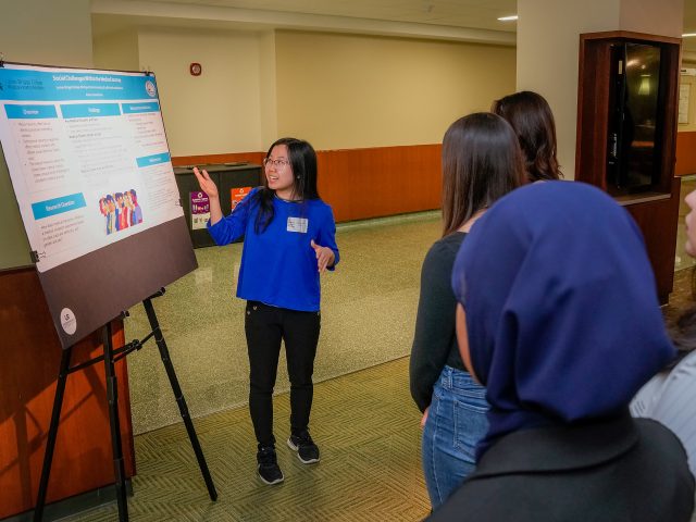 An Honors student presents research poster board to a group of people in the lounge of MSU Union, second floor.