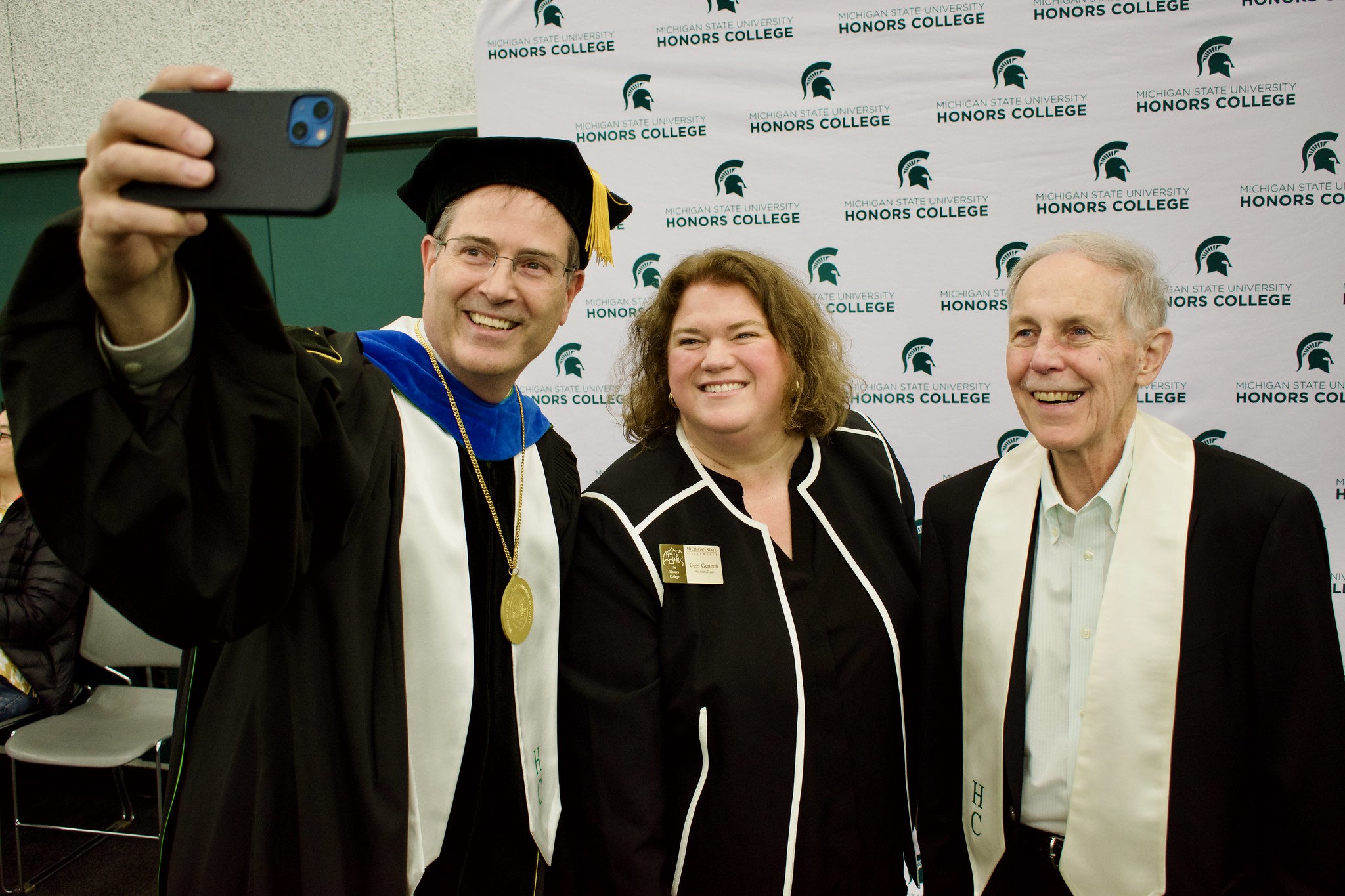 Chris Long, Bess German, and Ron Fisher pose for a selfie at a Michigan State University Honors College graduation reception. Long is wearing academic regalia, including a black graduation gown, cap, gold medallion, and white stole. German and Fisher are in black formal attire. A photo backdrop with the Honors College logo is visible in the background.