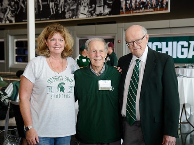 Molly Brennan, Ron Fisher, and M. Peter McPherson stand together at a Michigan State University event. Brennan is wearing a Michigan State Spartans t-shirt, Fisher is in a green sweater, and McPherson is dressed in a suit and tie. The setting includes a sports-themed backdrop and tables in the background.