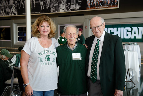 Molly Brennan, Ron Fisher, and M. Peter McPherson stand together at a Michigan State University event. Brennan is wearing a Michigan State Spartans t-shirt, Fisher is in a green sweater, and McPherson is dressed in a suit and tie. The setting includes a sports-themed backdrop and tables in the background.