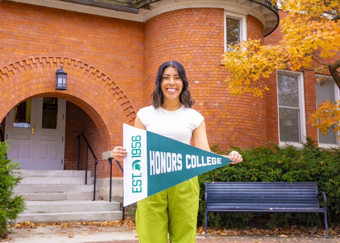 Jess Brandt holding a MSU Honors College pennant flag outside Eustace Cole Hall.