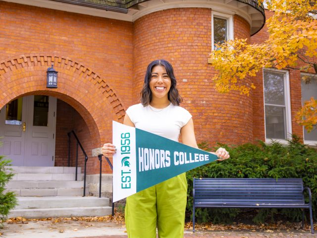 Jess Brandt holding a MSU Honors College pennant flag outside Eustace Cole Hall.