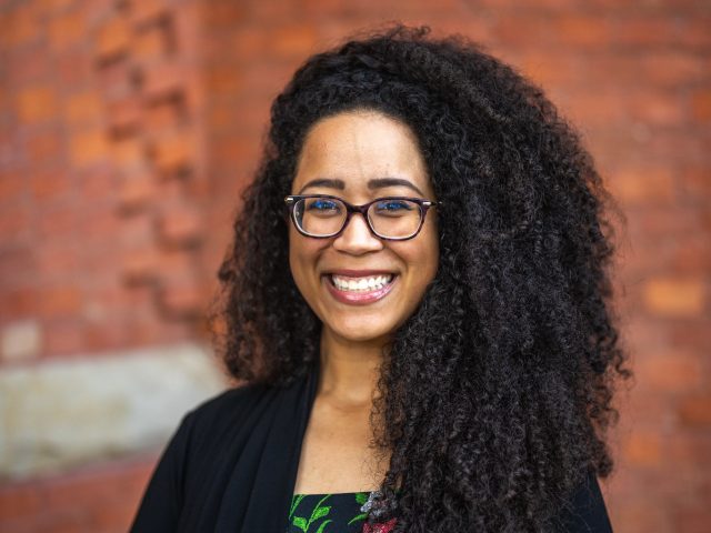 Image of Honors College DEIB Director Erika Trigg Crews smiling and standing in front of a red brick building. Crews is wearing a black cardigan and a woven floral shirt.