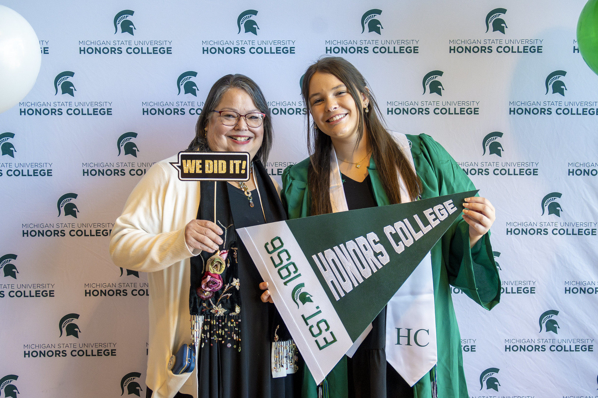 Image of Honors College student Brandi Stover in a green graduation gown and white collar stole, holding a green a white Honors College pennant. Next to Brandi is another person holding a small sign that says "WE DID IT!". Both people are smiling at the camera. Behind them is a white photo backdrop with a repeating pattern of green Spartan helmets and the words "MICHIGAN STATE UNIVERSITY HONORS COLLEGE".