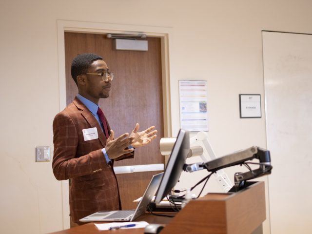 A student in a burnt orange suit, blue collared shirt, and red tie stands at a wooden podium, gesturing to the audience at the 2025 Diversity Research Showcase.