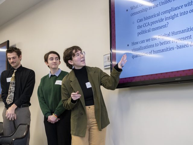 Three students stand at the front of the room, in front of two large screens with research slides on them. One student is looking at the screen and pointing, while the other two look on. All of them are dressed in business attire at the 2025 Diversity Research Showcase.