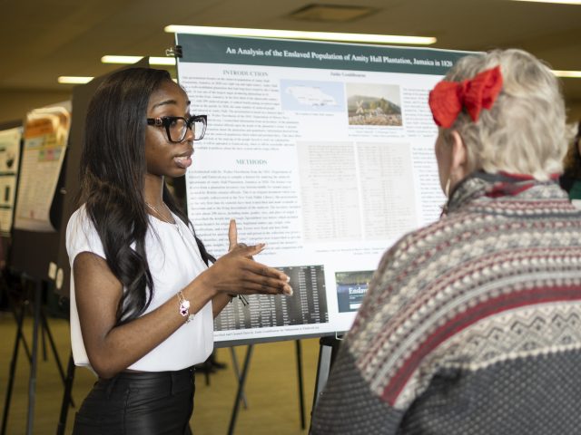 Honors College student Jaida Gouldbourne stands in front of a research poster, speaking with an engaged Diversity Research Showcase attendee.