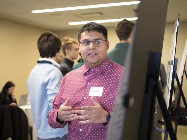 Honors College student Domenic Cedillo stands in front of a research poster at the Diversity Research Showcase. Cedillo is glancing at the poster and gesturing.