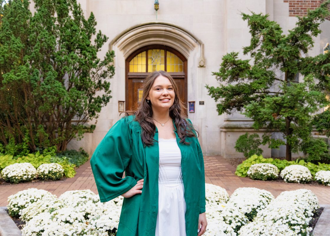 Image of Honors College student Brandi Stover standing with a hand on one and smiling at the camera. Brandi is wearing a green graduation gown and white dress. White flowers, a red brick path, green shrubs, and the tan stone of MSU's Beaumont Tower are in the background.